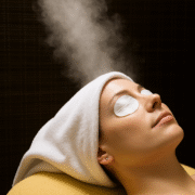 A woman lies peacefully on a spa bed with a white towel wrapped around her head and cotton pads over her eyes. Warm steam rises above her face, creating a soothing atmosphere. The background features a dark, textured surface, enhancing the calm and serene ambiance of the spa treatment.